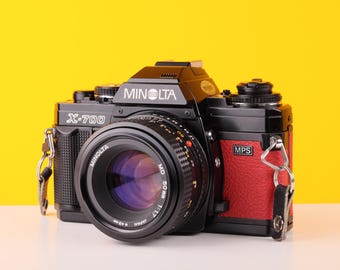 Pode incluir: Uma câmera de filme preta Minolta X-700 com uma empunhadura de couro vermelha e uma lente Minolta MD 50mm 1:1.7. A câmera tem uma alça prateada.