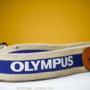 May include: Cream-coloured Olympus camera strap with a blue stripe and the brand name in white. The strap has a brown leather end piece with the Olympus logo. The background is a solid yellow.
