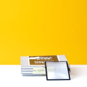 May include: A silver and black box with the text "Konica  Focusing Screen" printed on the top. A black focusing screen with a clear glass center is sitting on a white surface.