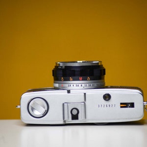 Puede incluir: Una c&aacute;mara de pel&iacute;cula de 35 mm plateada y negra con un objetivo que tiene un anillo negro con marcas para ajustes de apertura de 2,8 a 22. La c&aacute;mara tiene un cuerpo plateado con una correa de cuero negra.