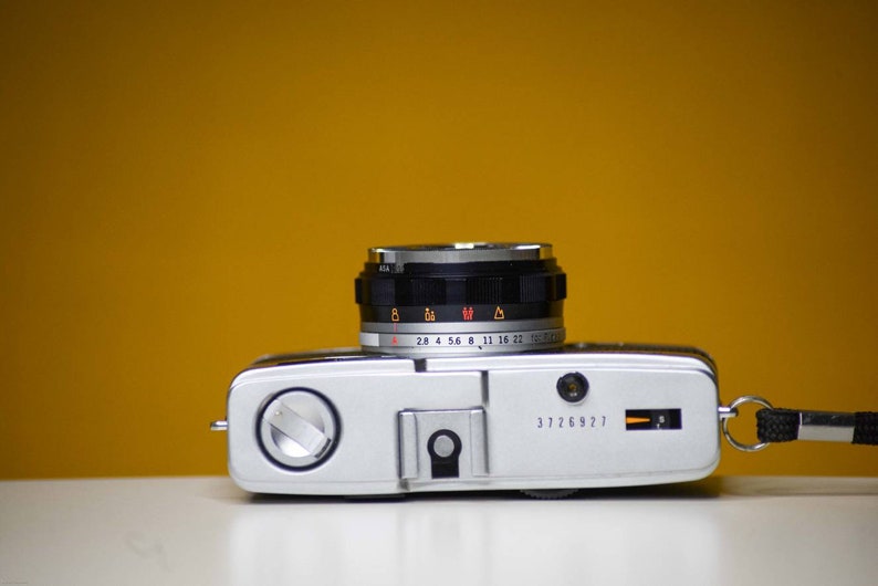 Puede incluir: Una c&aacute;mara de pel&iacute;cula de 35 mm plateada y negra con un objetivo que tiene un anillo negro con marcas para ajustes de apertura de 2,8 a 22. La c&aacute;mara tiene un cuerpo plateado con una correa de cuero negra.