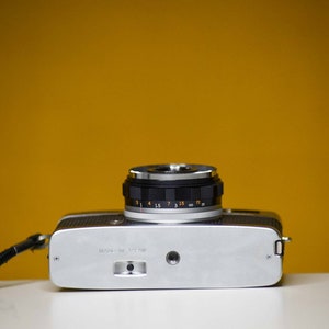 Puede incluir: Una c&aacute;mara de pel&iacute;cula de 35 mm de color plateado y negro con una lente negra y una correa. La c&aacute;mara tiene una escala de profundidad de campo en la lente.