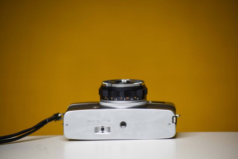 Puede incluir: Una c&aacute;mara de pel&iacute;cula de 35 mm de color plateado y negro con una lente negra y una correa. La c&aacute;mara tiene una escala de profundidad de campo en la lente.