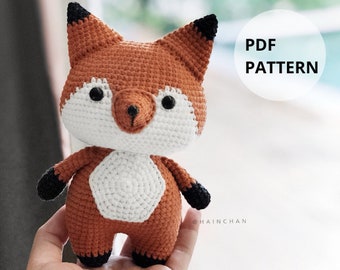 May include: Crocheted fox amigurumi toy with orange and white yarn. The fox has black eyes and a black nose. The toy is being held in a hand. PDF PATTERN.