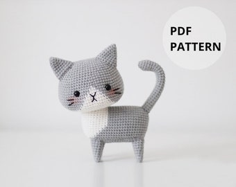 May include: A gray and white crocheted cat amigurumi toy with a white patch on its chest. The cat has black eyes and a pink nose. The image includes the text "PDF PATTERN".