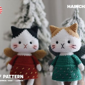 May include: Two crocheted cat angel toys with wings. The cat on the left is wearing a red dress with white polka dots. The cat on the right is wearing a green dress with white polka dots. The cats are both white with black and brown markings. The text "PDF PATTERN" and "HEIGHT APPROX: 17CM/ - 6.69INCH" is at the bottom of the image.