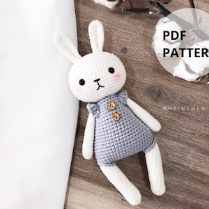 May include: A handmade white crochet bunny doll with a gray jumpsuit. The bunny has black eyes, pink cheeks, and a small black mouth. The jumpsuit has wooden buttons and ruffled sleeves. The text "PDF PATTERN" is in a circle.