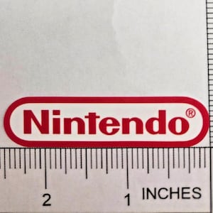May include: A red and white Nintendo logo sticker, approximately 5 cm long, with the word "Nintendo" in a bold, sans-serif font. The sticker is on a white background with an inch ruler for scale.