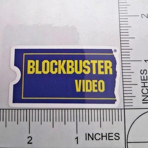 May include: A rectangular sticker featuring the Blockbuster Video logo. The sticker has a blue background with yellow text that reads "BLOCKBUSTER VIDEO." The sticker is approximately 5 cm long and 2.5 cm wide.