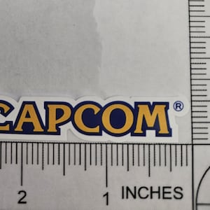 May include: A white sticker with the word "CAPCOM" in gold letters with a blue outline. The sticker is approximately 2 inches long and 0.5 inches tall. The sticker has a registered trademark symbol.
