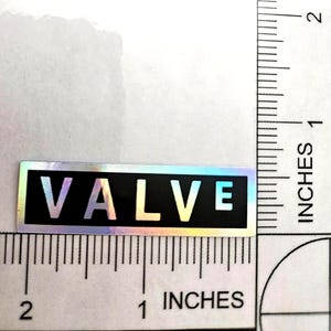 May include: A rectangular sticker with a black background and the word VALVE printed in white capital letters. The sticker has a holographic border and is positioned against a ruler showing it measures approximately 6.35 cm in length.