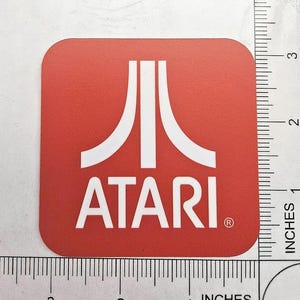May include: A square red coaster with rounded corners featuring the white Atari logo and text. A white ruler is positioned along the bottom and right edges, showing the coaster measures approximately 3.5 inches in width and height.
