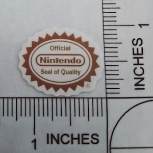 May include: A white and brown Nintendo Seal of Quality sticker. The sticker is oval-shaped with a brown border and the words "Official Nintendo Seal of Quality" printed on it. The sticker is approximately 2.5 cm in diameter.