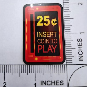 May include: A rectangular sticker with a red border and a black centre. The sticker reads "25¢" in yellow, with "INSERT COIN TO PLAY" below. The sticker is approximately 2.5 cm wide and 3.8 cm tall.
