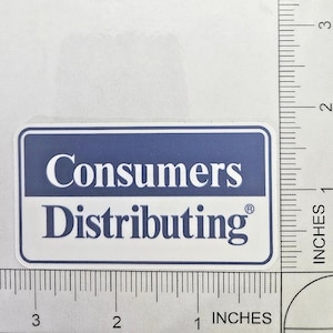 May include: A rectangular sticker featuring the Consumers Distributing logo with white text on a blue background and blue text on a white background. A ruler is visible along the side and bottom, showing the sticker is approximately 7.6 cm wide.