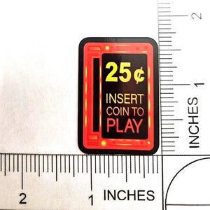 May include: A rectangular sticker with a black centre and a red border featuring the yellow text 25¢ and the white and orange text INSERT COIN TO PLAY. A ruler shows the sticker is approximately 3.8 cm tall and 2.5 cm wide.