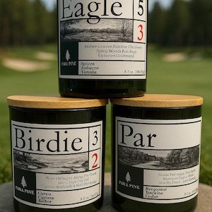 May include: Three green glass candles with wooden lids are stacked. Each candle has a label with a golf-themed name: "Eagle," "Birdie," and "Par." The labels include scent descriptions and the Park & Pine logo. Each candle is 8.5 oz.