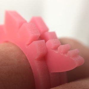 May include: A pink plastic ring with a textured, spiky design. The ring is worn on a finger.