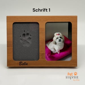Può includere: Una cornice in legno con due scomparti. Lo scomparto sinistro ha un'impronta di zampa grigia e il testo "Bella" inciso sotto. Lo scomparto destro contiene una foto di un piccolo cane bianco seduto su un letto rosa. La cornice ha il testo "Schrift 1" sopra.