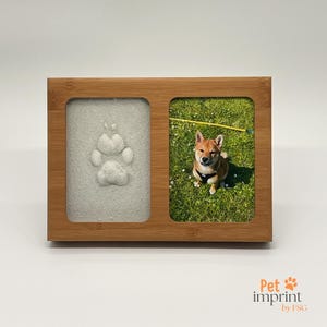 Può includere: Una cornice in legno con due scomparti, uno che mostra un'impronta di zampa bianca e l'altro una foto di un cane marrone e bianco seduto sull'erba verde. La cornice ha il testo "Pet imprint by FSG" sotto.