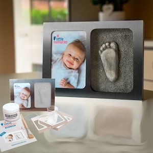 May include: A silver frame with a photo of a baby and a grey footprint cast in a grey frame. The frame is on a white surface with a white jar of plaster, a small brush, and a clear plastic tray with a grey square inside.