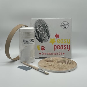Könnte beinhalten: Ein weißes Plastikgefäß mit Kopiergips, auf dem Etikett steht "COPY PLASTER ReliefgieBmasse". Das Gefäß steht neben einem Karton mit dem Text "easy peasy" und "Dein Abdruck in 3D" darauf. Auf einer weißen Oberfläche liegt eine weiße Gipsform mit einem Seestern und Muscheln.