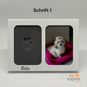 Può includere: Cornice bianca con un'impronta di zampa nera e una foto di un piccolo cane bianco con un fiocco rosa seduto su un letto rosa. La cornice ha il testo "Bella" e "Schrift 1" stampato su di essa.