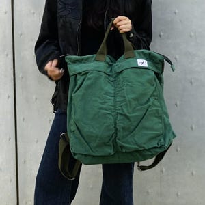 May include: A green corduroy tote bag with a top handle and shoulder straps. The bag has two front pockets and a small white tag with a logo. The bag is held by a person wearing a black jacket and blue jeans.