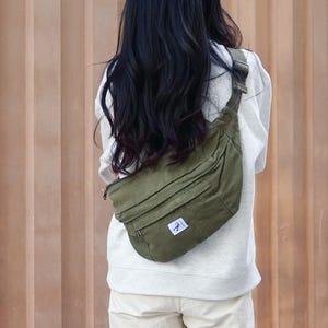 May include: An olive green bum bag with a small white logo patch. The bag is worn over the shoulder. The person is wearing a white sweatshirt and cream trousers. The background is a wooden wall.