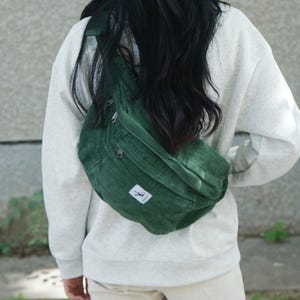 Może przedstawiać: Zielona nerka z sztruksu z małą białą etykietą. Torba ma główną przegrodę zapinaną na zamek i mniejszą przednią kieszeń. Torba jest noszona na ciele. Osoba ma na sobie szarą bluzę i beżowe spodnie.