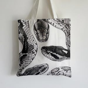 May include: White tote bag featuring a black and white illustration of several snake heads. The snakes are depicted in detail, with scales and patterns visible. The bag has white handles and is hanging against a white background.