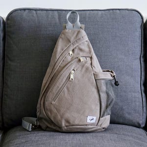 Peut inclure: Un sac bandoulière en toile beige avec plusieurs compartiments zippés et une poche latérale en filet. Le sac a une sangle réglable grise et un petit logo sur le devant. Il est posé sur un canapé gris.
