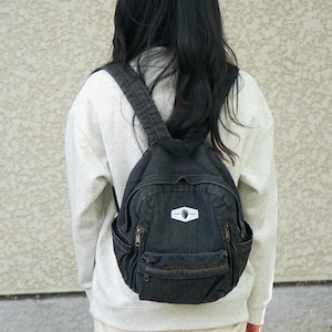 May include: A black denim backpack with multiple zippered compartments and adjustable straps. The backpack features a small white patch with a logo. The bag is worn on the back.