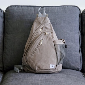 May include: A tan canvas sling bag with multiple zipped compartments and a mesh side pocket. The bag has a grey adjustable strap and a small logo on the front. It is on a grey sofa.