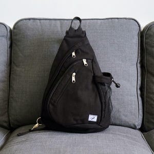 Può includere: Zaino a tracolla nero con più scomparti con cerniera e una tasca laterale in rete. La borsa ha una maniglia superiore e una piccola etichetta bianca con un logo. La borsa è realizzata in un tessuto resistente e ha una cerniera dorata.