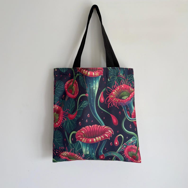 May include: A tote bag with a dark blue background and a repeating pattern of red and teal pitcher plants. The bag has black handles and is hanging against a white wall. The plants have a detailed, stylized design.