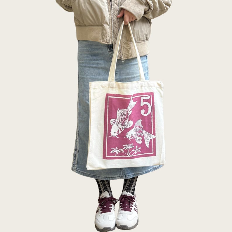 May include: A white canvas tote bag featuring a magenta rectangular design with two koi fish and the number 5. The tote bag has long handles and is held by a person.