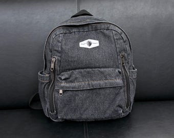 Pode incluir: Uma mochila de ganga cinzento-escura com pega superior, alças ajustáveis e um bolso frontal com fecho de correr. O saco inclui dois bolsos laterais e um emblema retangular branco na parte superior com um pequeno gráfico e texto ilegível.