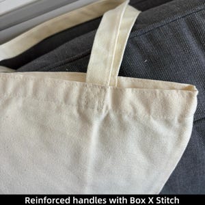 May include: Close-up of a cream-colored tote bag with reinforced handles. The handles are attached with a box X stitch. The bag is made of a sturdy, textured fabric. The text "Reinforced handles with Box X Stitch" is at the bottom of the image.