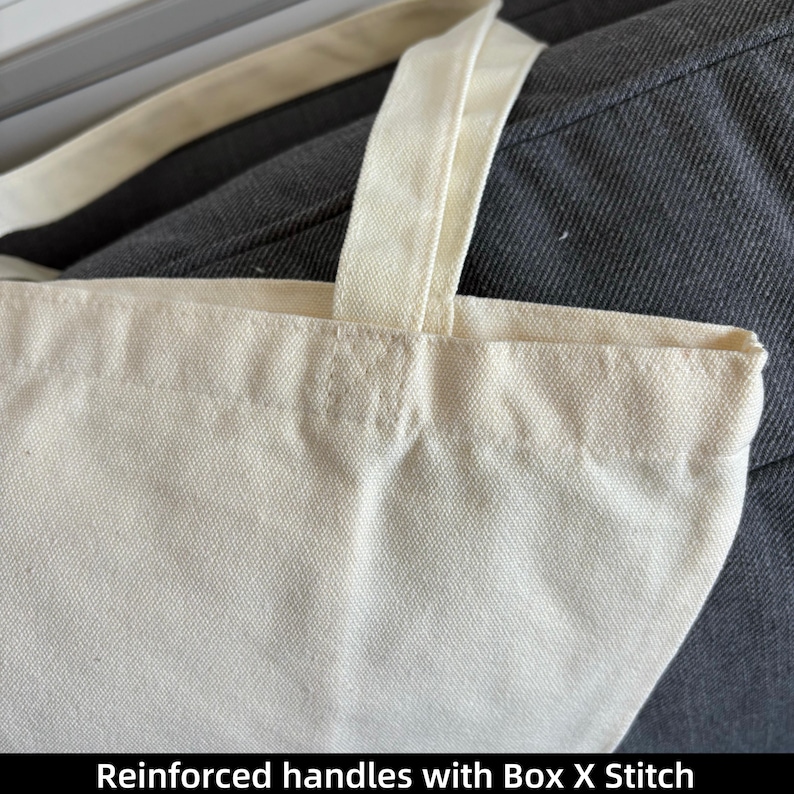 May include: Close-up of a cream-colored tote bag with reinforced handles. The handles are attached with a box X stitch. The bag is made of a sturdy, textured fabric. The text "Reinforced handles with Box X Stitch" is at the bottom of the image.