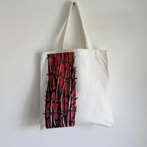 May include: White tote bag with a striking graphic. The design features a vertical red rectangle with black tree-like shapes and barbed wire. The bag has long white handles and is hanging against a white wall.