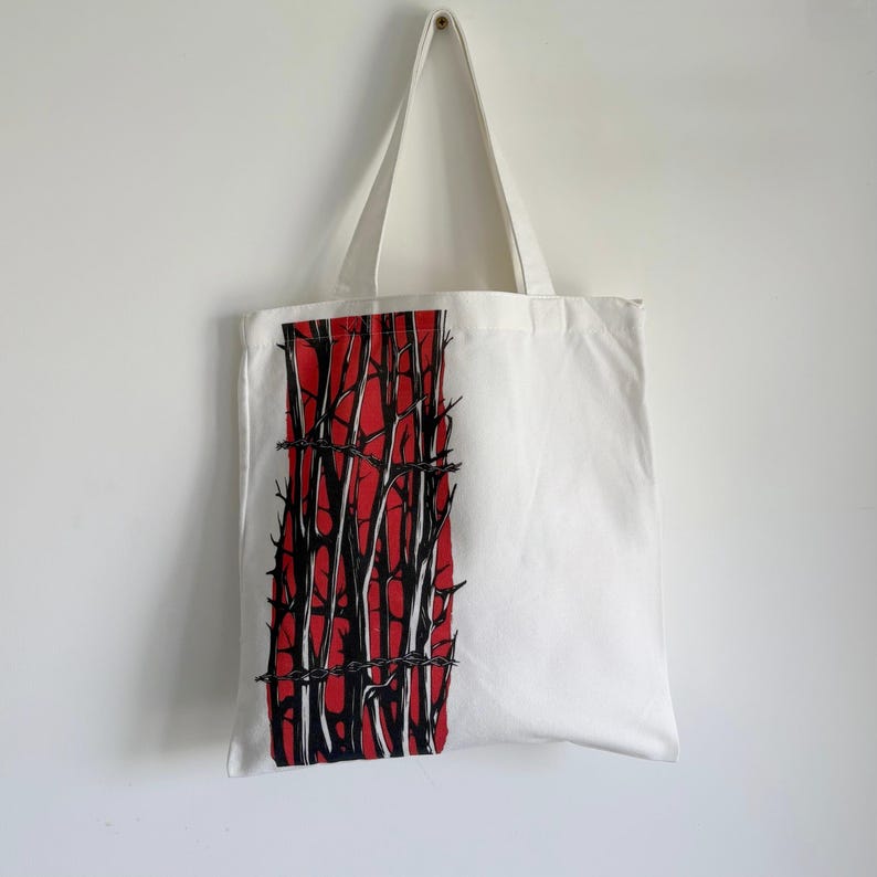 May include: White tote bag with a striking graphic. The design features a vertical red rectangle with black tree-like shapes and barbed wire. The bag has long white handles and is hanging against a white wall.