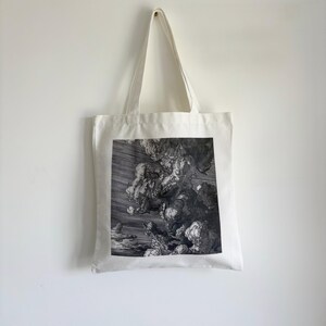 May include: A white tote bag with a black and white print of a dramatic cloudscape. The image features swirling clouds and a dark, stormy sky. The bag has long, white handles and is hanging against a white wall.