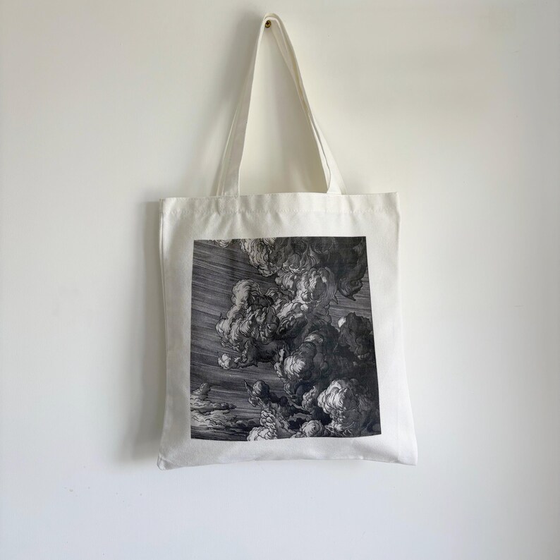 May include: A white tote bag with a black and white print of a dramatic cloudscape. The image features swirling clouds and a dark, stormy sky. The bag has long, white handles and is hanging against a white wall.