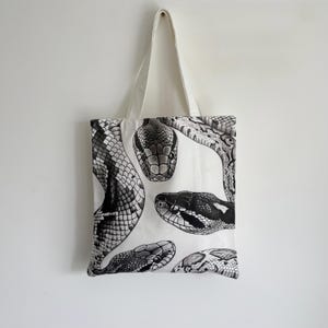 May include: White tote bag featuring a black and white illustration of several snake heads. The snakes are depicted in detail, with scales and patterns visible. The bag has a simple design with a white handle.