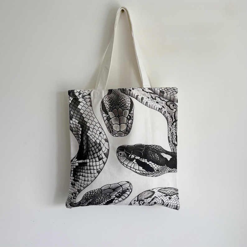 May include: White tote bag featuring a black and white illustration of several snake heads. The snakes are depicted in detail, with scales and patterns visible. The bag has a simple design with a white handle.