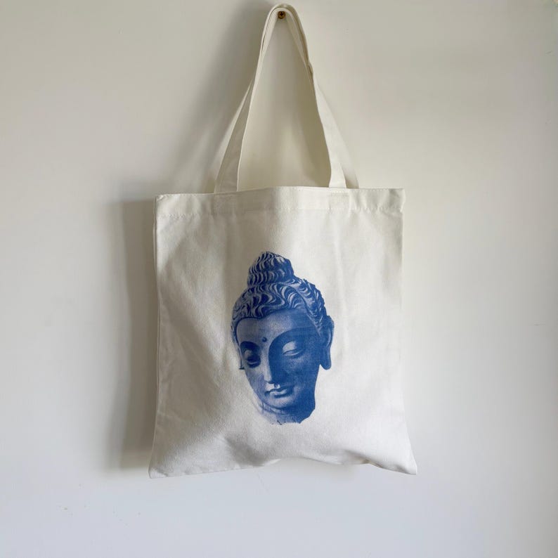 May include: A cream-colored tote bag with a blue graphic of a Buddha's head. The tote bag has two long handles and is hanging against a white wall. The Buddha's head is centered on the bag and is a detailed illustration.