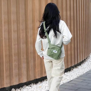 Pode incluir: Uma pessoa vestindo uma camisa branca de manga comprida e calças cáqui, carregando uma bolsa transversal verde. A bolsa tem um pequeno patch branco com um design preto. O fundo apresenta uma parede de madeira e uma passarela pavimentada.