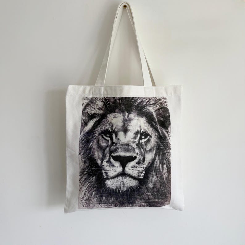 May include: White tote bag featuring a detailed black and white illustration of a lion's face. The tote bag has two white handles and is hanging against a white background. The lion's face is centered on the bag.
