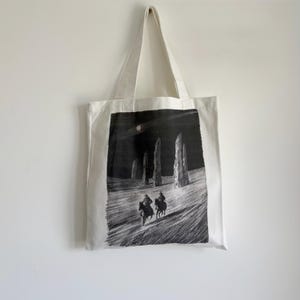May include: White canvas tote bag featuring a black and white illustration of two riders on horseback in a landscape with tall, stone structures under a night sky. The bag has a rectangular design with a handle.